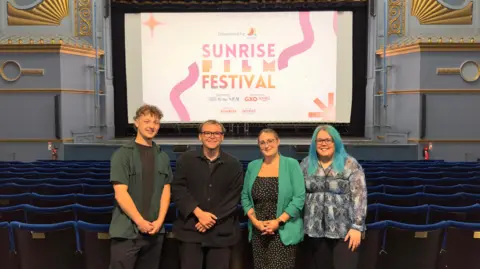 SUNRISE STUDIOS Four people are standing beside each other in front of theatre stage showing sign for Sunrise Film Festival