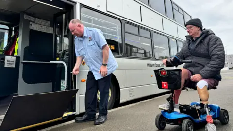 Eric Corkish is sitting on his blue mobility scooter on the pavement. He is wearing a hat and black coat, and shorts. His left left is a prosthetic. A bus driver is pulling down a ramp on to the pavement from the doorway of a silver double-decker bus.