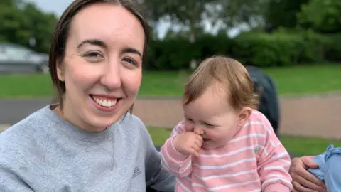 Johanna Patterson has long dark hair tied back and is wearing a grey top and holding a baby with a pink and white striped top. The baby has her hand in her mouth.