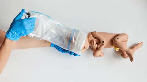 The OdonAssist being used on a baby replica doll. A person wearing blue gloves is using the device which has a plastic handle with a soft plastic extension which is sitting around the baby's head.