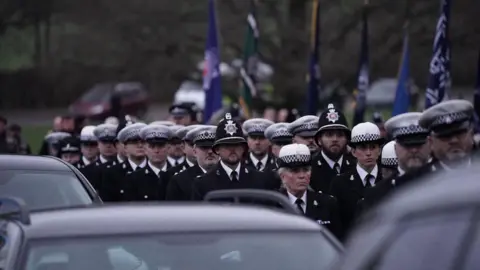 Essex Police A long line of police officers wearing suits and hats. They are walking in line as part of a funeral procession. A row of black cars are next to them, as are various flags being held aloft.