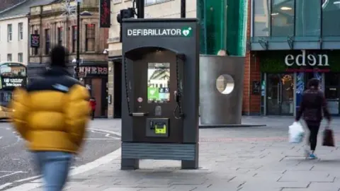 JCDecaux/In Focus Networks Ltd A high street showing a row of shops and a communication hub in the middle of the pavement. It looks like a dark grey phone box and says "defibrillator" on the top. The blurry figures of two people are walking past