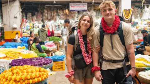 Contestants Sioned and Fin are in a busy and colourful market Indian market. They are both wearing backpacks.