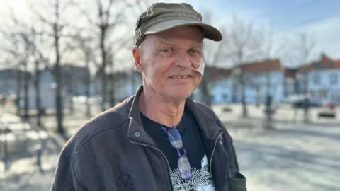 BBC A man with a cap on his head and a medical tube coming from his nose smiles at the camera