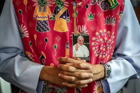 LUIS TATO / AFP A Catholic nun prays during a Requiem Mass in honour of Pope Francis. She wears a red, patterned tunic and pinned to her clothing is a portrait of the late Pope.
