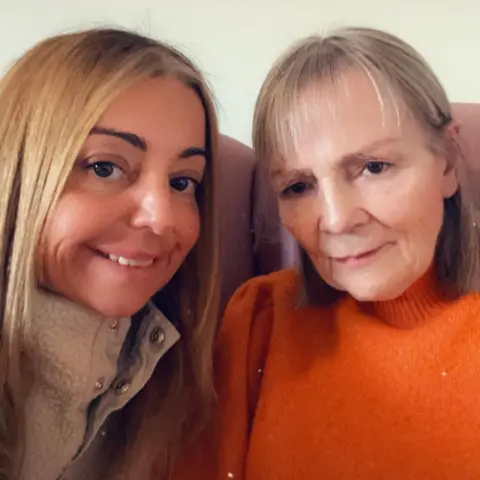 Nicola Hughes selfie Nicola dengan ibunya Janette. Nicola mengenakan bulu krem ​​dan Janette mengenakan jumper oranye. Mereka duduk di sofa merah muda.