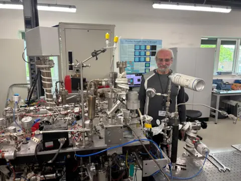 BBC News Uma grande rede de tubos, fios de metal, cabos de plástico em uma mesa em um laboratório. Sasha Verchovsky fica atrás da máquina, chamada Finesse.