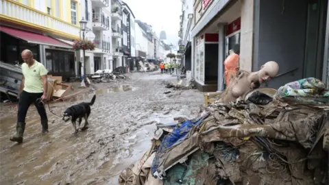 EPA A person wearing rubber boots walks with a dog on a leash near debris sitting along a street after flooding in Bad Neuenahr-Ahrweiler, Germany, 16 July 2021