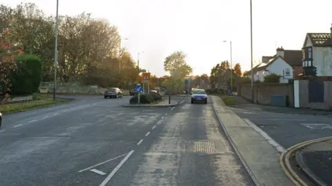 A wide residential road with traffic on each side. Houses, street signs and trees can be seen in the background.