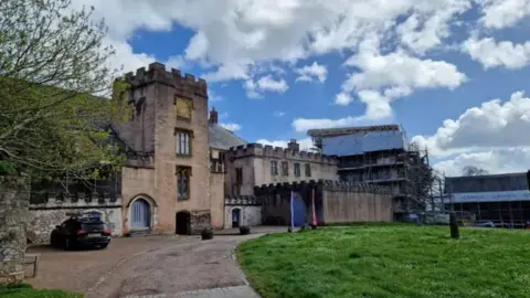 The photo shows branches of a tree on the left-hand side which has green leaves. Next to it is Torre Abbey, which has turrets of a castle in the middle and along the roof of the building. There is a green space on the right-hand side of the photo which is part of the golf course.