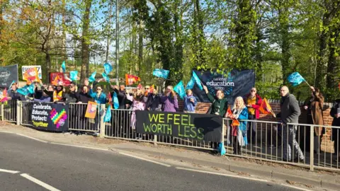 Dozens of people waving blue flags stand at the side of a road in a protest. There are banners reading 'we feel worth less' and advertising the NEU.