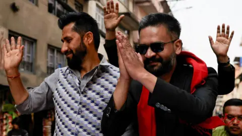 Reuters Rabi Lamichhane, head of the Rastriya Swatantra Party, wears a patterned grey and blue shirt and waves at a crowd next to Balendra Shah who wears sunglasses, a black suit and red scarf and whose palms are clasped in front of his face
