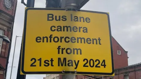 BBC Close up of a yellow street sign with black writing on a lamppost saying 'bus lane camera enforcement from 21st May 2024'. There are red brick buildings in the background and a grey, cloudy sky