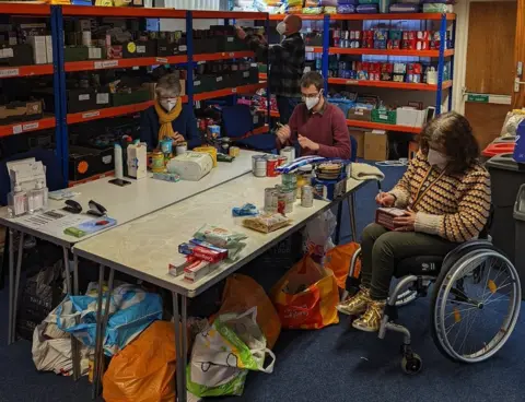 People at a table with food items stacked on shelves by them