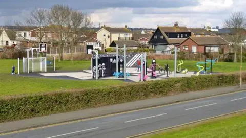 Kismet Park, which is next to a road and surrounded by houses and a green bush. The park has various equipment including a football goal, basketball nets and an outdoor gym.