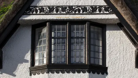 Historic England Archive A zoomed in view of one of the windows of a building which has a thatched roof. The frame of the window is painted brown, and there is pattern above the window. 