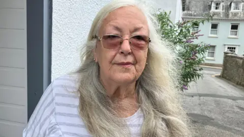 Norma Cattell, with glasses and long grey hair, standing in front of the community larder