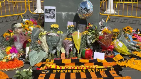 Matthew Cooper/PA Wire Floral tributes along with scarves, photographs and football T-shirt are on the ground with yellow metal barriers behind.