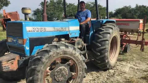 Ronald sits on a blue tractor in his farm.