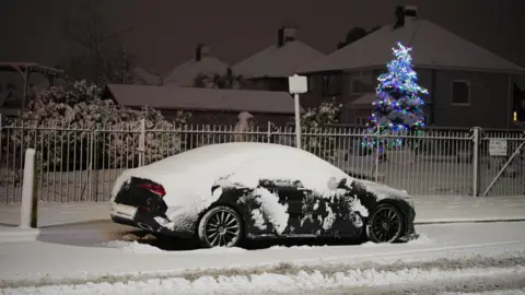 PA Media A black car covered in snow parked at the side of the road. The ground and houses behind the car are covered in snow. A tree decorated with Christmas lights can be seen in the garden behind the car.