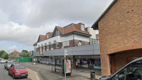 The outside of Lidl supermarket in Horley which is a shop below what look like homes about with a mix of red brick and white plaster. The windows of the supermarket have writing and pictures of food on them and there are cars parked outside and a bus stop.