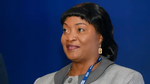 Jewel Cianeh Taylor wears a blue lanyard and grey jacket, gold earring at an economic forum in Poland, 2021. She has shiny black hair.