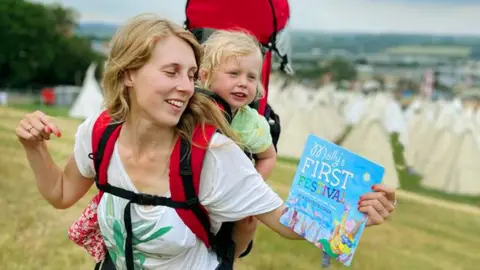 Katherine O'Shea A young blonde haired woman with a young toddler in a baby carrier on her back