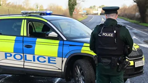 A police man, with his back to the camera, stood on a road with a police car beside him. There are rows of houses and greenery in background.
