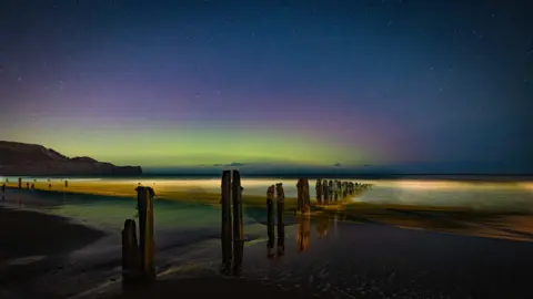 The Aurora Borealis - known as the Northern Lights - over Sandsend in the North York Moors National Park. The lights are shining green, purple and dark blue while the sea appears to be glowing a golden colour.