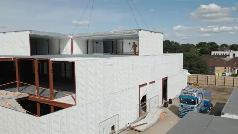 A half-build white building with workmen stood on it, and a lorry to the right of it