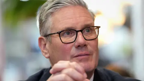 Sir Keir Starmer looks away from camera while wearing glasses and clasping his hands against a bokeh background, as he visits a kitchen in Kensal Rise on 18 May.