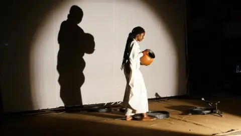 Latifatou Ouédraogo pours water from a jug into barrels to open the premiere the first act of Là-bas ou Ici (Here or There) at the Institut Français in Ouagadougou on April 23, 2021.