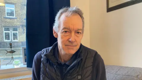 A man wearing a black gilet and zip-up jumper underneath sitting inside a house