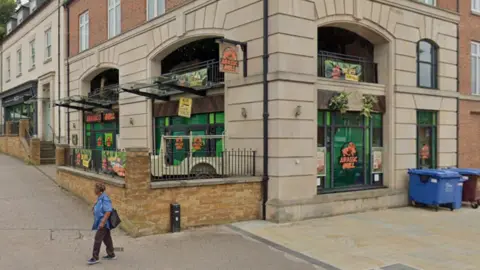 The outside of a restaurant called Jurassic Grill. A woman is walking past. There is a large blue bin on the right and green signs in the windows. There is a wall with railings in front of the building.