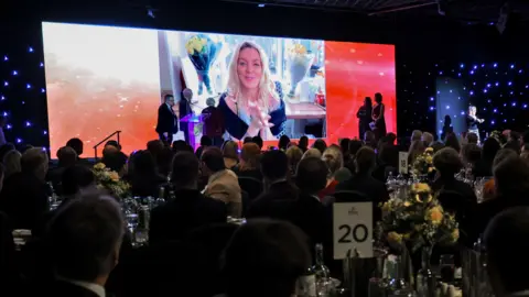 North News and Pictures Sheridan Smith appears on a large screen above a stage. Peopple sitting at tables watch on.