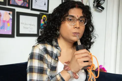 A woman with curly long dark hair and clear rimmed glasses puffs on a large cannabis vaporiser with an orange lead trailing out of it