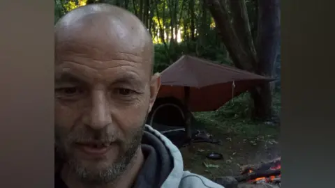 Blair Murray has a shaved head with a stubbly beard. He is wearing a grey hoodie and stands in front of a fire and tent. His tent has a tarpaulin strung over his tent in the middle of woodland. The picture is a selfie.