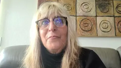 Susan, a woman with long blonde hair and half-rimmed glasses, sitting on a leather sofa with some abstract art behind her, on a video call with the BBC