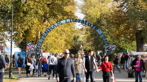 A sparse crowd of people walk through a park down a wide pavement lined by trees. There is a blue arch over the pavement which reads "Cheltenham Literature Festival".