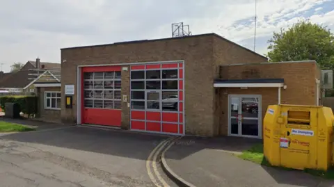An external view of a brick-built fire station. It has two large red painted doors with glass windows and a lower building attached, with a doorway. 