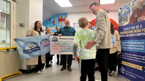 Harry (left) and father (Edward) are seen laughing as Bluebell Ward staff hold a giant check and smile at them. 