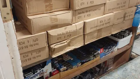 Lots of cardboard boxes stacked in a backroom. On the bottom shelves are packets of cigarettes and vapes. 