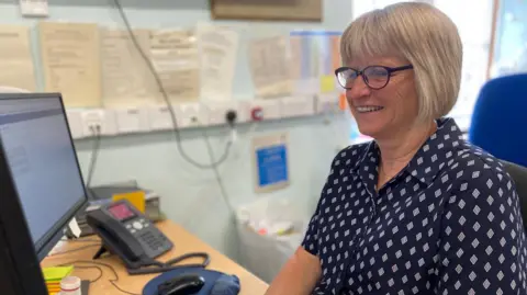 A woman wearing a patterned navy blouse sits in an office smiling at a computer. She has short blonde hair and wears black glasses.