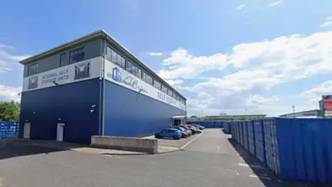 An existing storage yard owned by O'Brien on Ferryboat Lane. A large blue industrial office building stands to the left. There is a row of cars parked outside. Around three sides of the building are rows of blue storage containers.