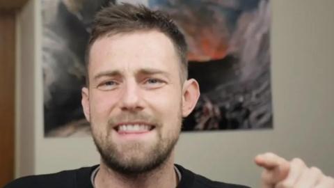 Joshua Sutcliffe, in a still taken from his YouTube channel, has a short beard, short dark hair, and is pointing at the camera. A picture, which appears to show a volcano erupting and/or a Biblical scene, is behind him on a white wall