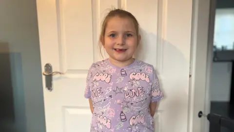 Eight-year-old Phoebe in a purple top stands smiling against a white door in her living room