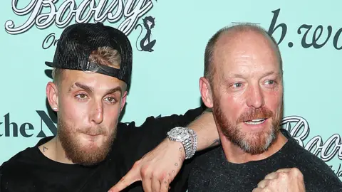 Getty Images Jake Paul and his father Greg Paul looking away from the camera. The background is light blue with branding, Jake is wearing a cap backwards and a silver watch on his left hand, which is resting on Greg's right shoulder. They are both wearing dark tops.