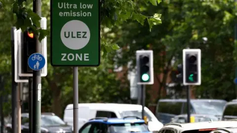 Reuters File image of stationary cars at traffic lights below a green Ulez sign.