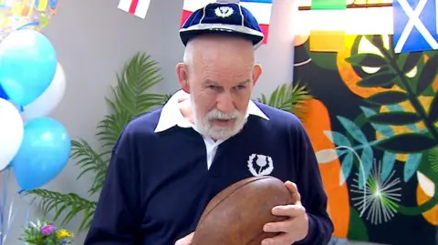 An older man in a navy rugby top and cap holding a rugby ball