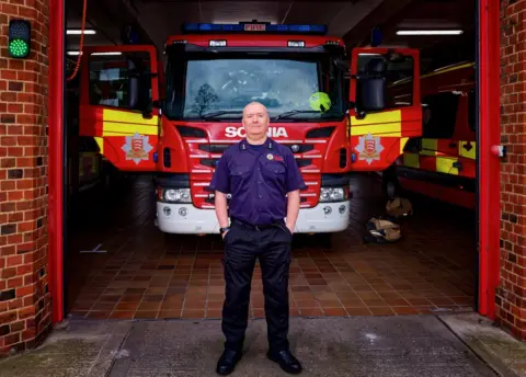 Terry Maher in front of Colchester fire station with a fire engine behind him